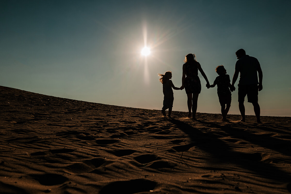 Family of four silhouetted holding hands on sand dunes at sunset during West Michigan family photography session GAUPERfamilies