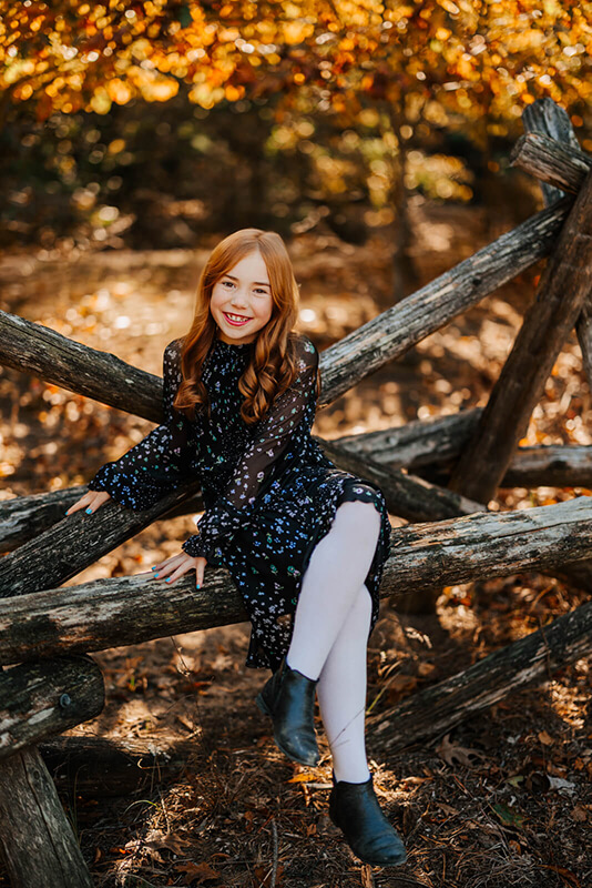 Young girl with red hair sitting on a log fence smiling during fall family photography session Grand Haven Michigan