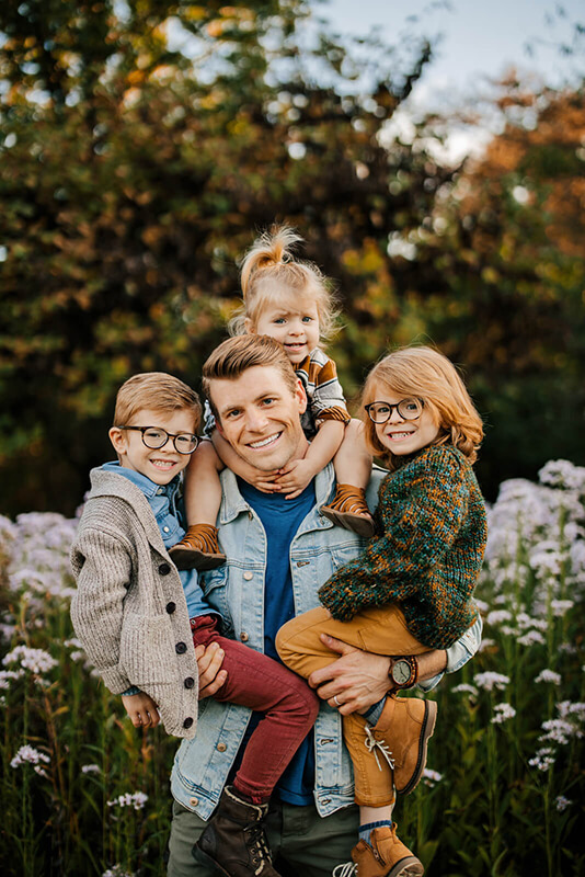 Dad with three kids piled on him laughing in wildflower field during Grand Rapids family photography session GAUPERfamilies