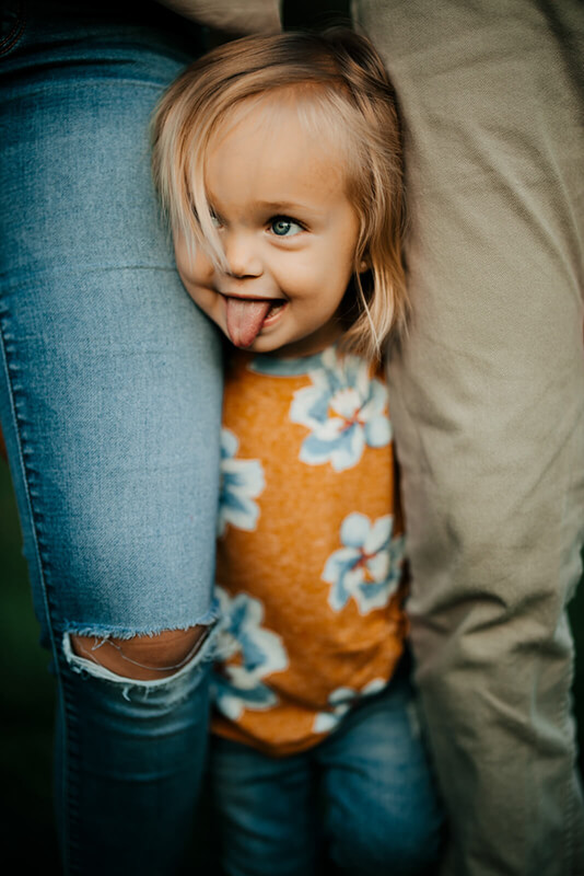 Toddler peeking between parents legs sticking out tongue during candid family photography session Muskegon Michigan