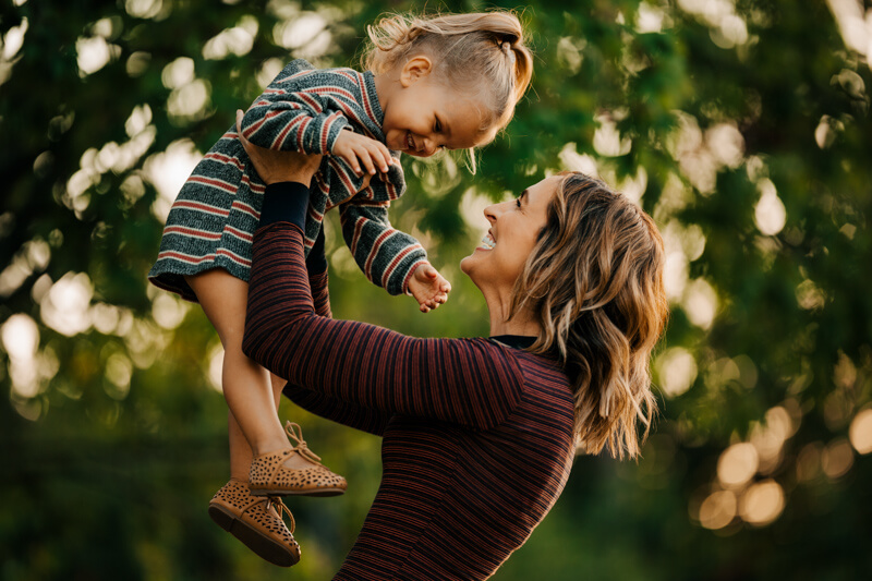 Mother lifting laughing toddler in the air during golden hour candid family photography session Muskegon Michigan